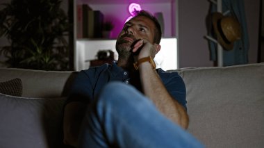 Düşünceli, Latin kökenli, gri saçlı, evdeki loş ışıklı oturma odasında oturan olgun bir adam.