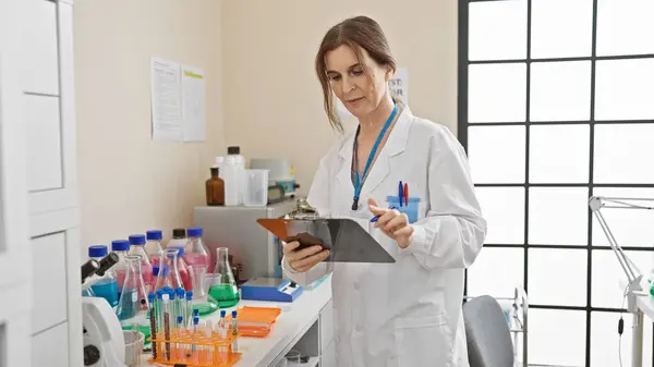 stock image Mature woman scientist analyzing clipboard in laboratory with equipment and window backdrop