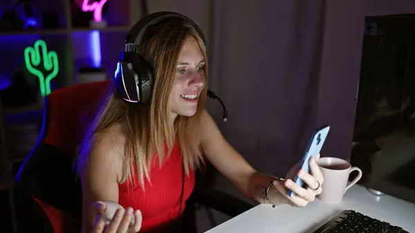 stock image Smiling woman with headphones using smartphone in a neon-lit gaming room at night