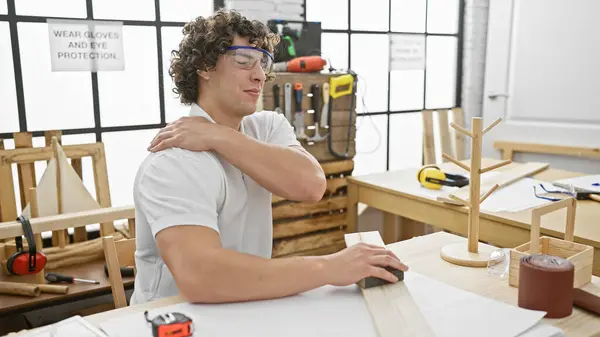 Stock image Young hispanic man with curly hair wearing safety glasses experiences shoulder pain in a well-lit carpentry workshop.