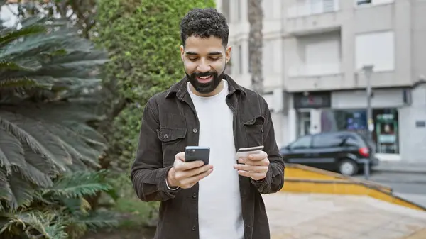 stock image A young hispanic man holding a credit card and smartphone smiles while standing outdoors in a city park.