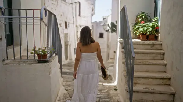 stock image A young hispanic woman in a white dress walks through the charming old town of ostuni in puglia, italy, surrounded by whitewashed buildings and potted plants.