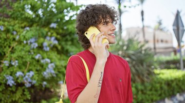 Kıvırcık saçlı genç bir adam sarı bir telefonla konuşuyor. Şehir parkında, gündüz vakti..