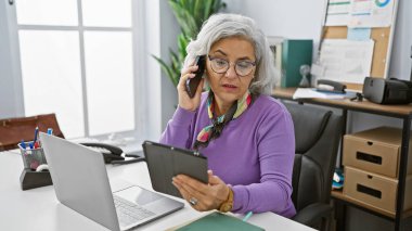Modern bir ofis ortamında telefon ve tabletle birden fazla iş yapan olgun bir kadın..
