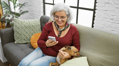 Neşeli, olgun bir kadın, evinde rahat bir kanepede köpeğine sarılırken akıllı telefonunu kullanmaktan zevk alır..