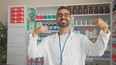 Gülümseyen erkek eczacı, ilaç rafları ve sağlık ürünleriyle donatılmış iyi stoklanmış bir eczanede kendini işaret ediyor..