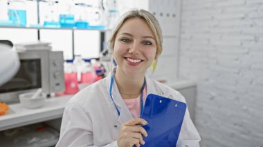 Beyaz önlüklü, gülümseyen genç bir kadın modern bir laboratuvar ortamında elinde bir pano tutuyor.