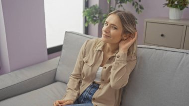 Genç ve çekici sarışın bir kadın, evinde oturma odasında otururken eli kulağında bir jest yapıyor..