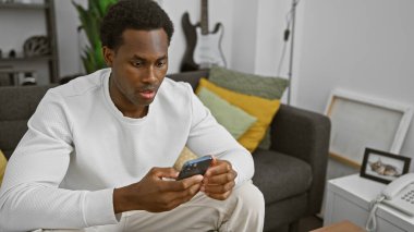 Modern bir oturma odasında akıllı telefon kullanan odaklanmış genç bir zenci.