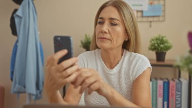 Orta yaşlı beyaz bir kadın, iyi aydınlatılmış bir oturma odasında akıllı telefon kullanır. Düşüncelerini ya da endişelerini dile getirir..
