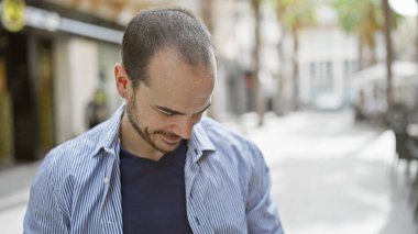 Çizgili tişörtlü, gülümseyen kel bir adam güneşli bir şehir sokağında duruyor ve sakin bir şehir ortamını canlandırıyor..