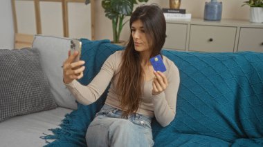 Genç bir İspanyol kadın oturma odasında elinde kredi kartıyla oturmuş evde selfie çekiyor ve sıradan bir ev dekoru sergiliyor..