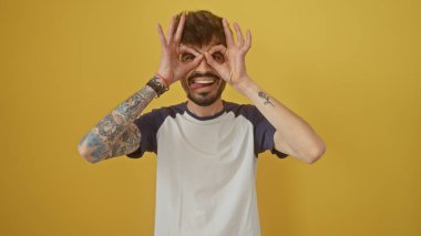 Dövmeli neşeli bir genç adam sarı bir arka plana karşı gözlük hareketi yapıyor..