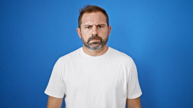 Beyaz tişörtlü olgun İspanyol bir adam. Dışarının mavi arka planına karşı. Ciddi bir ifadeyi resmediyor..