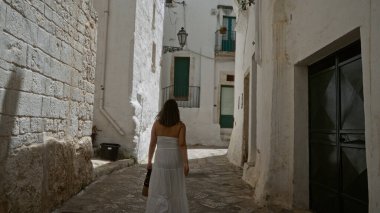 Beyaz elbiseli genç bir İspanyol kadın, geleneksel bembeyaz binaları ve dar kaldırımlı kaldırımlı caddeleriyle eski güzel Ottuni, Puglia, İtalya 'yı keşfediyor..