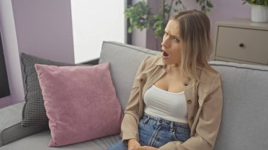 A beautiful young adult blonde caucasian woman in a stylish outfit expressing surprise while sitting on a couch in a cozy living room of a modern apartment. clipart