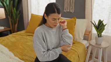 Genç bir İspanyol kadın, rahat bir yatak odasında, gri bir kazak giyerken dirseğini tutarak acı içinde yatağında oturuyor..