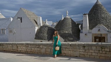 Yeşil elbiseli güzel İspanyol bir kadın dramatik bir gökyüzünün altındaki eşsiz Trulli evlerini hayranlıkla seyrederek eski Alberobello, İtalya 'da bir kaldırım taşı caddesinde duruyor..