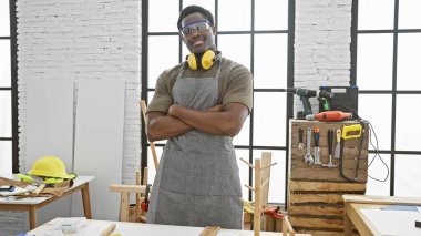 İyi organize edilmiş bir marangozluk atölyesinde kolları çapraz duran kendinden emin Afro-Amerikan bir adam..