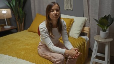 Güzel bir genç kadın, evdeki şık dekore edilmiş bir yatak odasında, rahat bir yatakta, modern iç dekorasyon ve iç mekan bitkileriyle birlikte, rahat bir şekilde oturur..