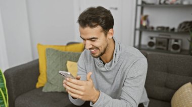Yakışıklı İspanyol adam rahat oturma odasında telefona gülümsüyor..