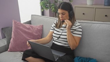 Odaklanmış bir kadın, rahat oturma odasından bilgisayarı üzerinde çalışıyor ve kulaklıkla bir telefon görüşmesi yapıyor..