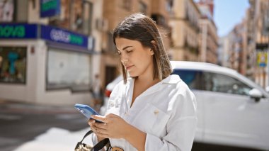 Genç ve güzel İspanyol kadın şehirde akıllı telefon kullanarak mutlu bir şekilde gülümsüyor.