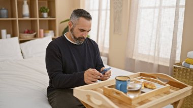 Gri sakallı, olgun bir İspanyol adam iyi ışıklandırılmış bir yatak odasında akıllı telefon kullanıyor, kahvaltı tepsisinin yanında oturuyor..