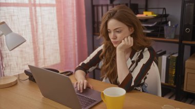 Genç ve çekici sarışın bir kadın ev ofisindeki bir masada dizüstü bilgisayarda çalışıyor. Raflar, belgeler ve arka planda görünen sarı bir bardak var..