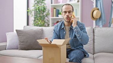 Bald hispanic man with beard talking on phone in a modern living room, perplexed by contents of a carton box. clipart