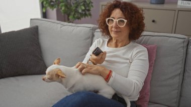 Orta yaşlı bir kadın oturma odasında elinde uzaktan kumandayla bir chihuahua ile kanepede oturuyor..