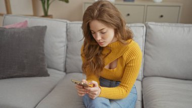 Sarışın, güzel bir kadın zarif dekore edilmiş bir oturma odasında otururken akıllı telefonunu kullanıyor. Huzurlu ve rahat bir ev ortamını canlandırıyor..