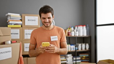 Gülümseyen genç bir İspanyol, kapalı bir bağış merkezinde gönüllü olarak çalışıyor, kutuların yanında akıllı telefon kullanıyor..