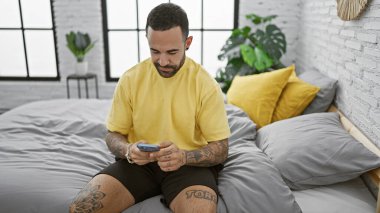 Sarı tişörtlü, sakallı genç bir adam gri bir yatakta oturuyor. Kapalı bir yerde akıllı telefon kullanıyor. Yatak odasının atmosferi sakin..