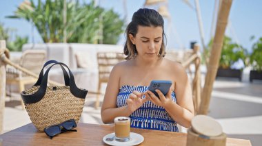 Genç İspanyol kadın akıllı telefon kullanıyor güneşli restoran terasında masada oturuyor.