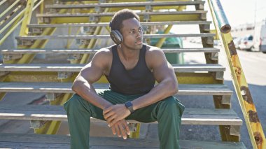 Kulaklık takan genç bir adam şehir ortamında dışarıda oturuyor, düşünceli görünüyor..