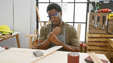 Afro-Amerikalı acı çekiyor. Marangozluk atölyesinde bileğini tutuyor. Güvenlik gözlükleri takılı..