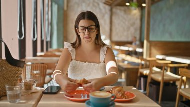 Lezzetli bir kruvasanın tadını çıkaran güzel bir İspanyol kadının portresi. Şu gözlüklü esmeri görüyor musun? Güneşli bir kafeterya masasında bu tatlı yemeğin tadını çıkarıyor. Kahvaltıyı ev içi bir zevk yapıyor..