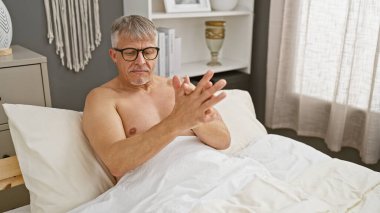 Tişörtsüz, orta yaşlı, gözlüklü ve gri saçlı bir adam iyi aydınlatılmış bir yatak odasında acı içinde kıvranıyor..