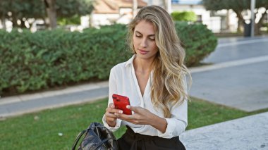 Havalı ama ciddi, genç sarışın bir kadın akıllı telefonunun dünyasına dalmış, bankta oturmuş güneşli park sükunetinin tadını çıkarıyor..