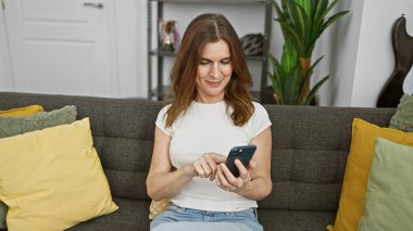 Modern bir oturma odasındaki koltukta akıllı telefon kullanan orta yaşlı bir kadın..