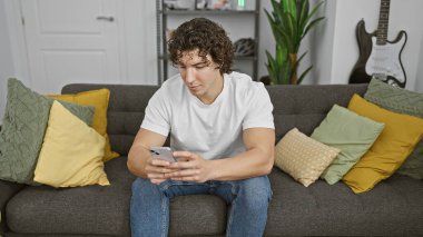 Modern oturma odasındaki koltukta akıllı telefon kullanan kıvırcık saçlı İspanyol adam..