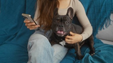 Genç bir İspanyol kadın oturma odasında elinde cep telefonuyla oturuyor. Yanında da mavi kanepede oturan köpeğine eşlik ediyor..