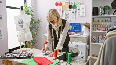 Sarışın bir kadın terzi, kumaş, iplik ve dikiş makinesiyle çevrili iyi organize edilmiş atölyesinde dikkatlice çalışıyor..