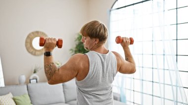 Sağlığını ve yaşam tarzını ifade eden, parlak bir oturma odasında dambıl kullanan, dövmeli, zinde bir genç adam..