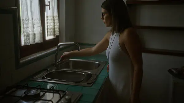 stock image Young hispanic woman in a typical old italian house, standing by the sink in a dimly lit kitchen with vintage decor.