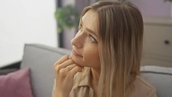 stock image Attractive young blonde caucasian woman in a home living room, contemplating with her chin resting on her hand.