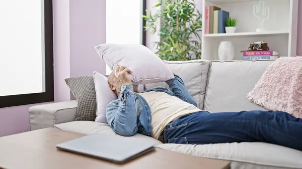 stock image A mature man with grey hair lies on a couch indoors, covering his face with a pillow in a relaxed home environment.