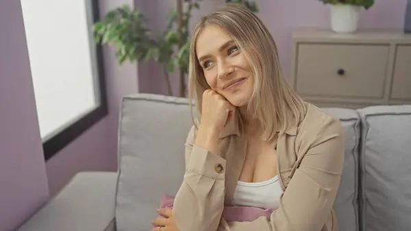 stock image A beautiful young caucasian woman sits in her living room, smiling thoughtfully while leaning on her hand, with a cozy home interior in the background featuring plants and soft furnishings.