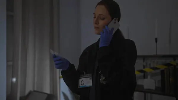 stock image A woman detective in gloves with a badge and evidence at a dark indoor crime scene, talking on the phone.